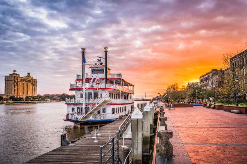 A classic paddle boat is moored at the docks in Savannah, GA with a beautiful sunset in the background.