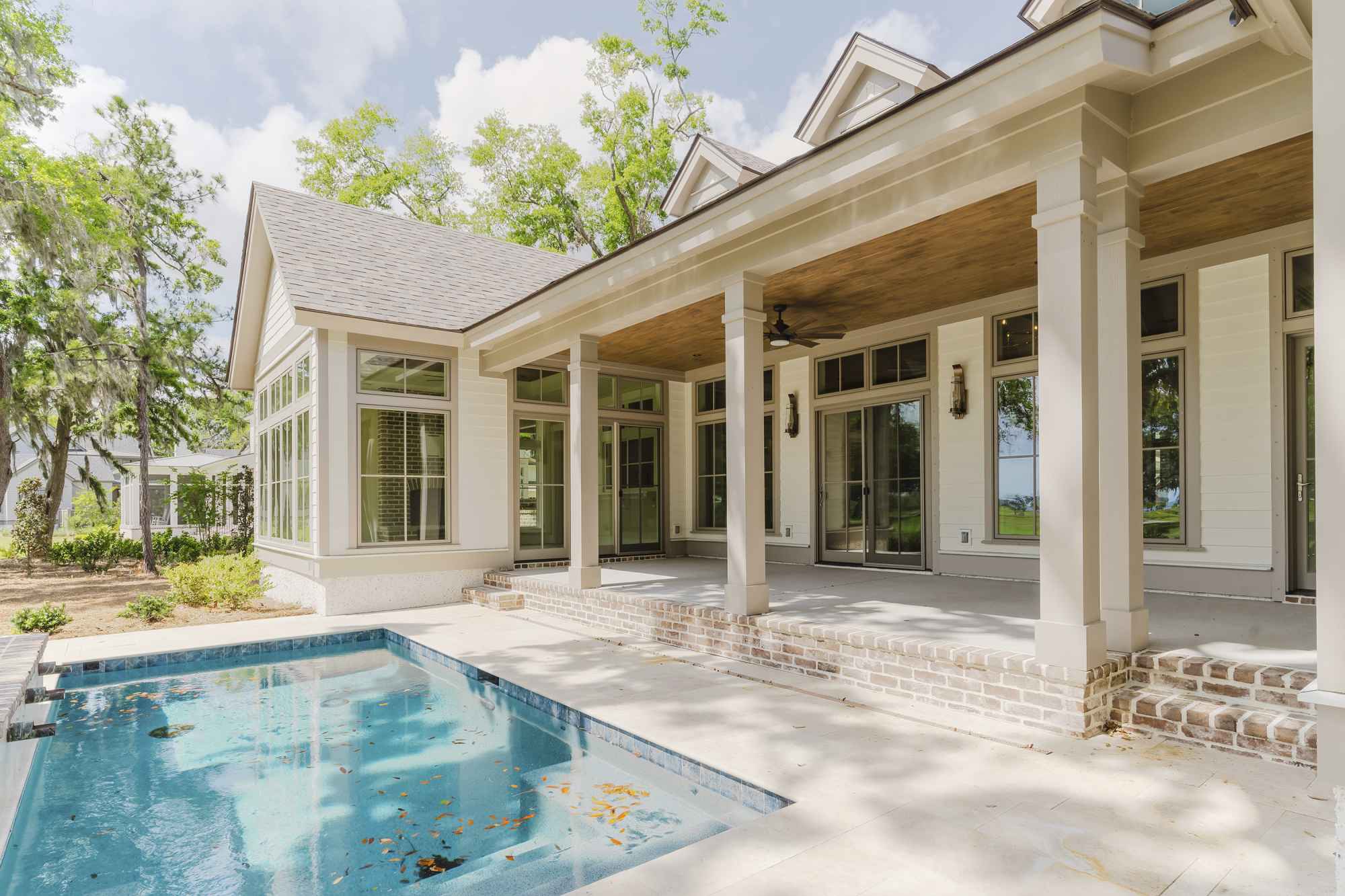  spacious modern house with large windows and a covered patio overlooks a small, leaf-strewn pool. Surrounded by lush green trees on a sunny day.