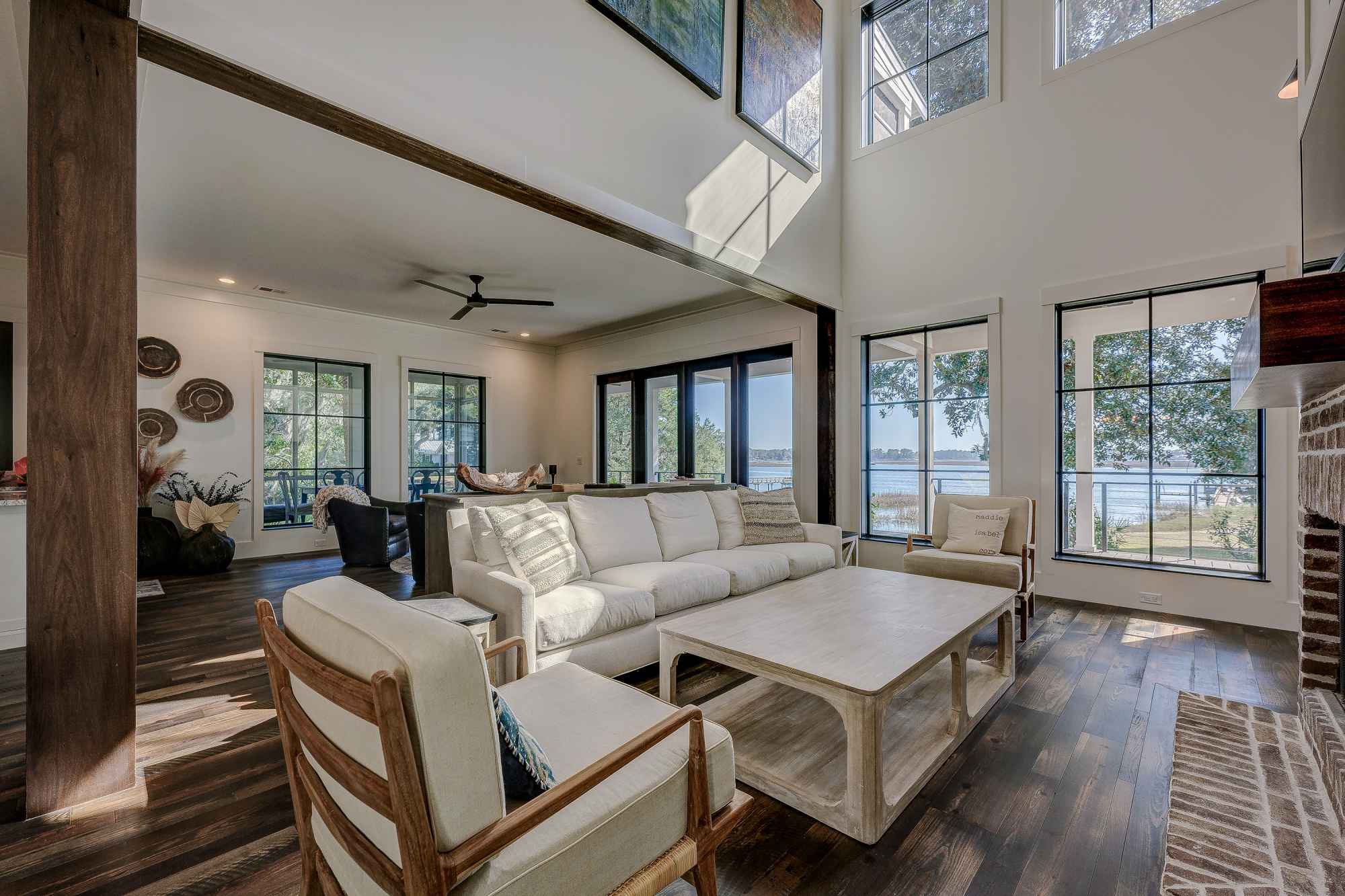 Sunny living room with high ceilings features a beige sofa set, wooden floors, large windows with lake views, and a modern ceiling fan. Airy and serene atmosphere.