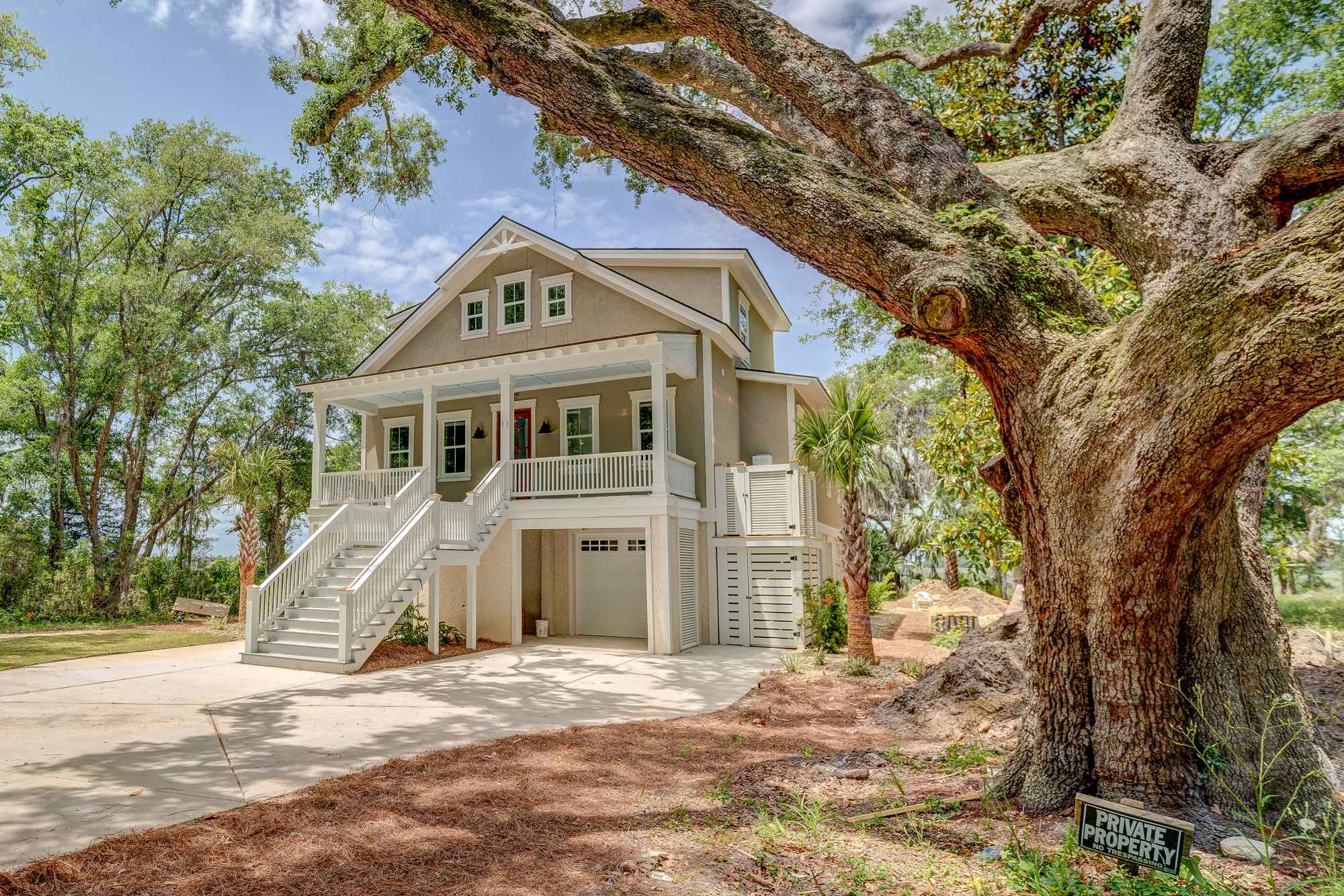 A charming two-story house with a raised porch is surrounded by lush trees. A large, old oak tree stands prominently in the foreground.
