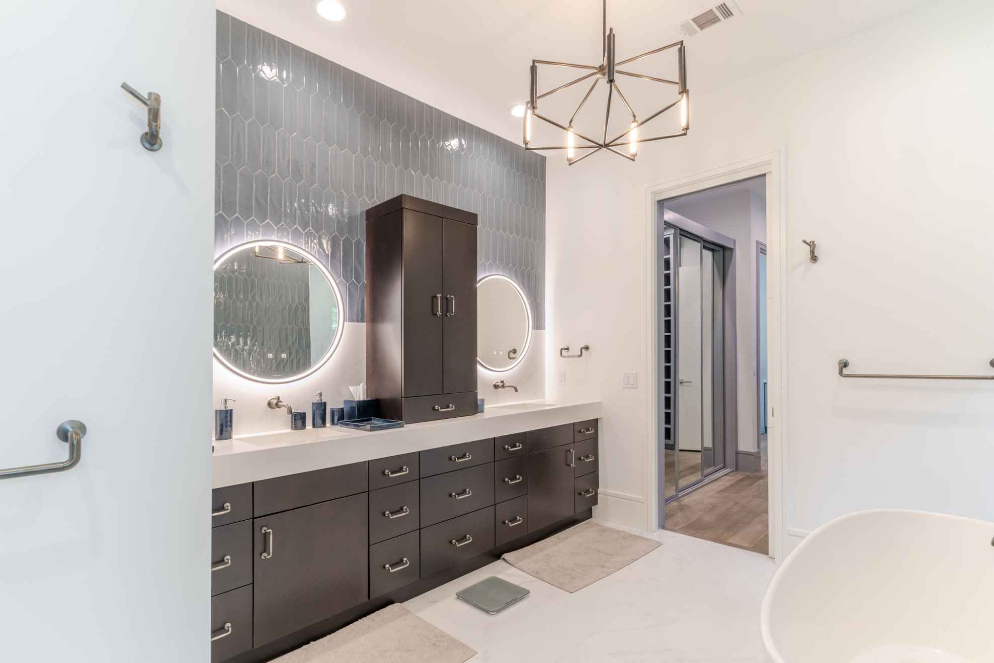 A modern bathroom featuring dual sinks with illuminated circular mirrors, dark cabinetry, a geometric light fixture, and a soothing, elegant decor.