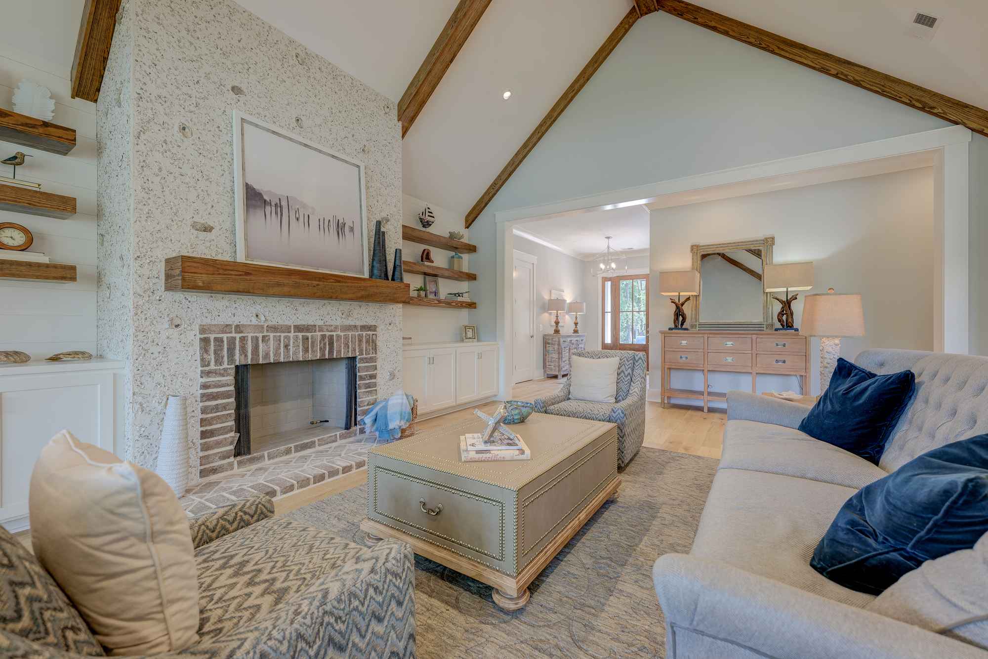 Cozy living room with high, wooden beam ceilings, a stone fireplace, plush beige sofas, blue cushions, and rustic wooden shelves.