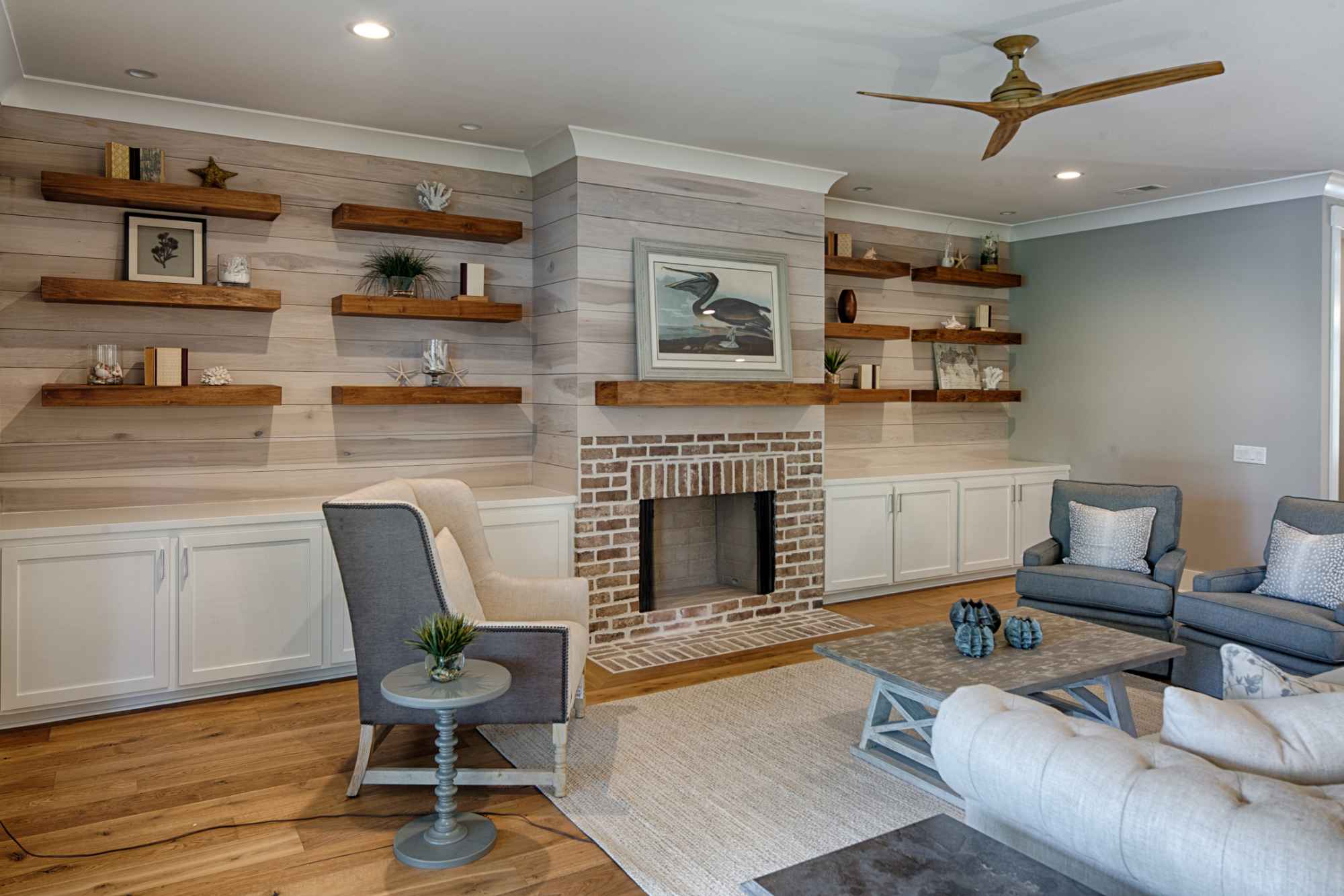 Cozy living room with a brick fireplace, flanked by wooden shelves displaying decor with neutral tones, cushioned seating, and a ceiling fan. 