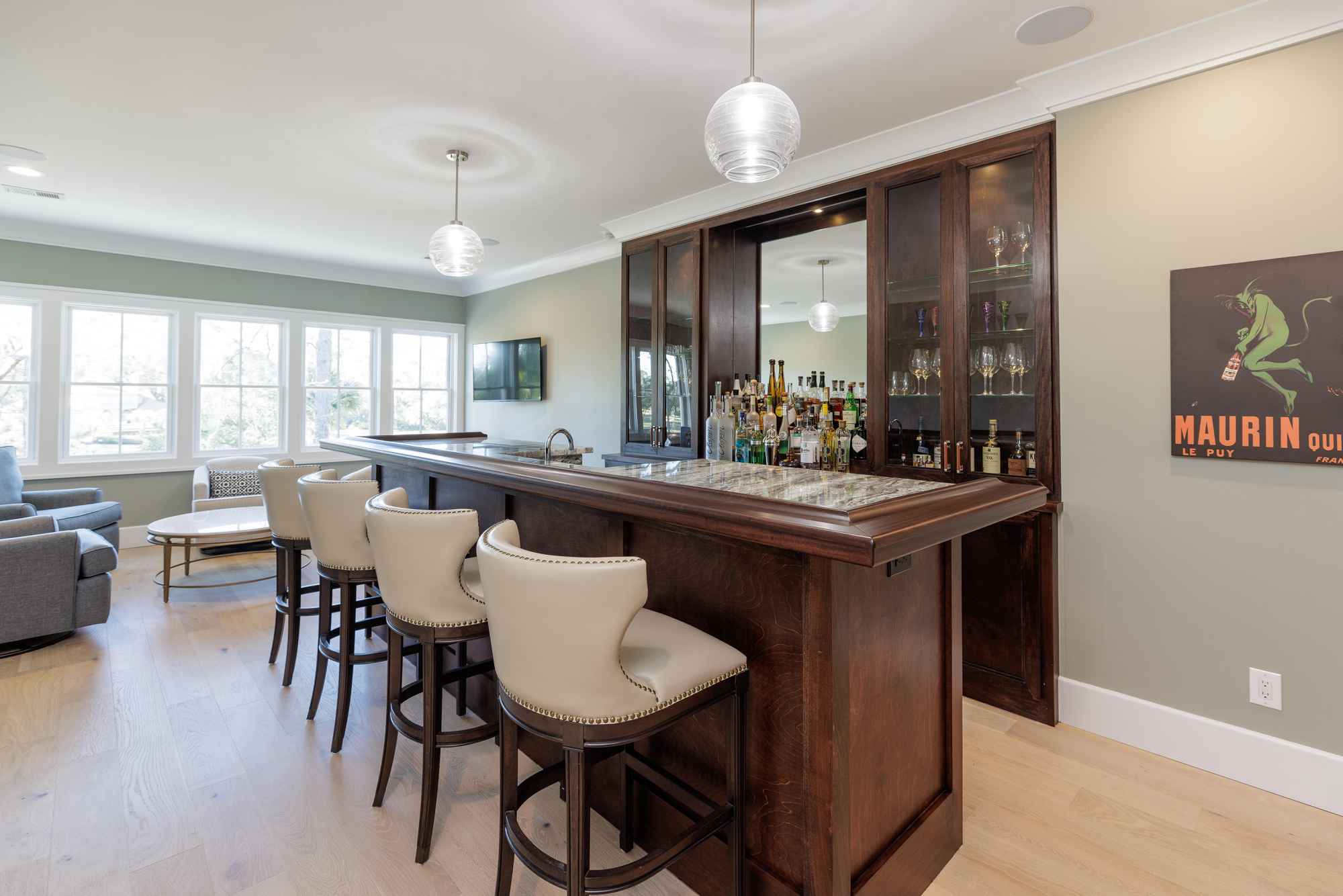 A stylish home bar area features a dark wood counter with stools, a mirrored back bar with glass cabinets filled with bottles and glassware. The room is well-lit and has a modern yet comfortable aesthetic.