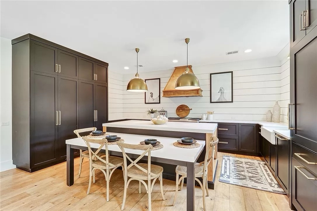 A modern kitchen with a farmhouse style features dark cabinets, a large island with wooden chairs, shiplap walls, gold pendant lights, and wood flooring.