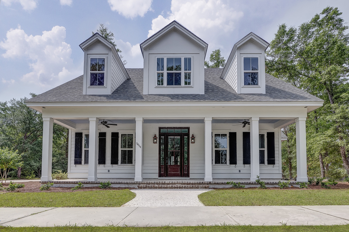 White two-story house with symmetrical gables, a covered porch, and black shutters. Surrounded by trees and under a blue sky with fluffy clouds.