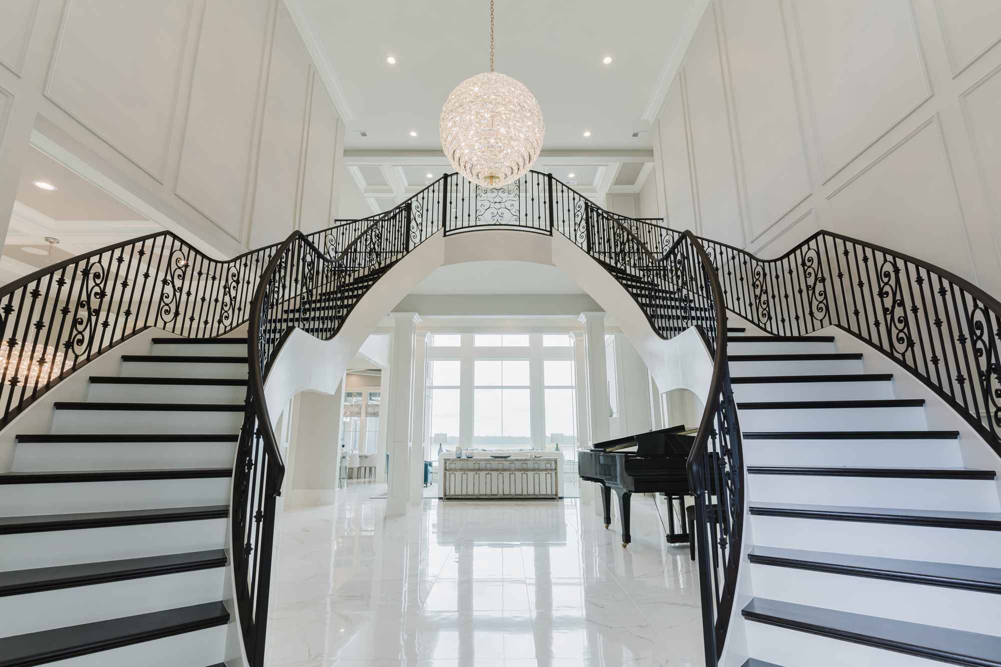 A classic double staircase rises from the white marble tile floor. Between the two sets of stairs large floor-to-ceiling great room windows are revealed.