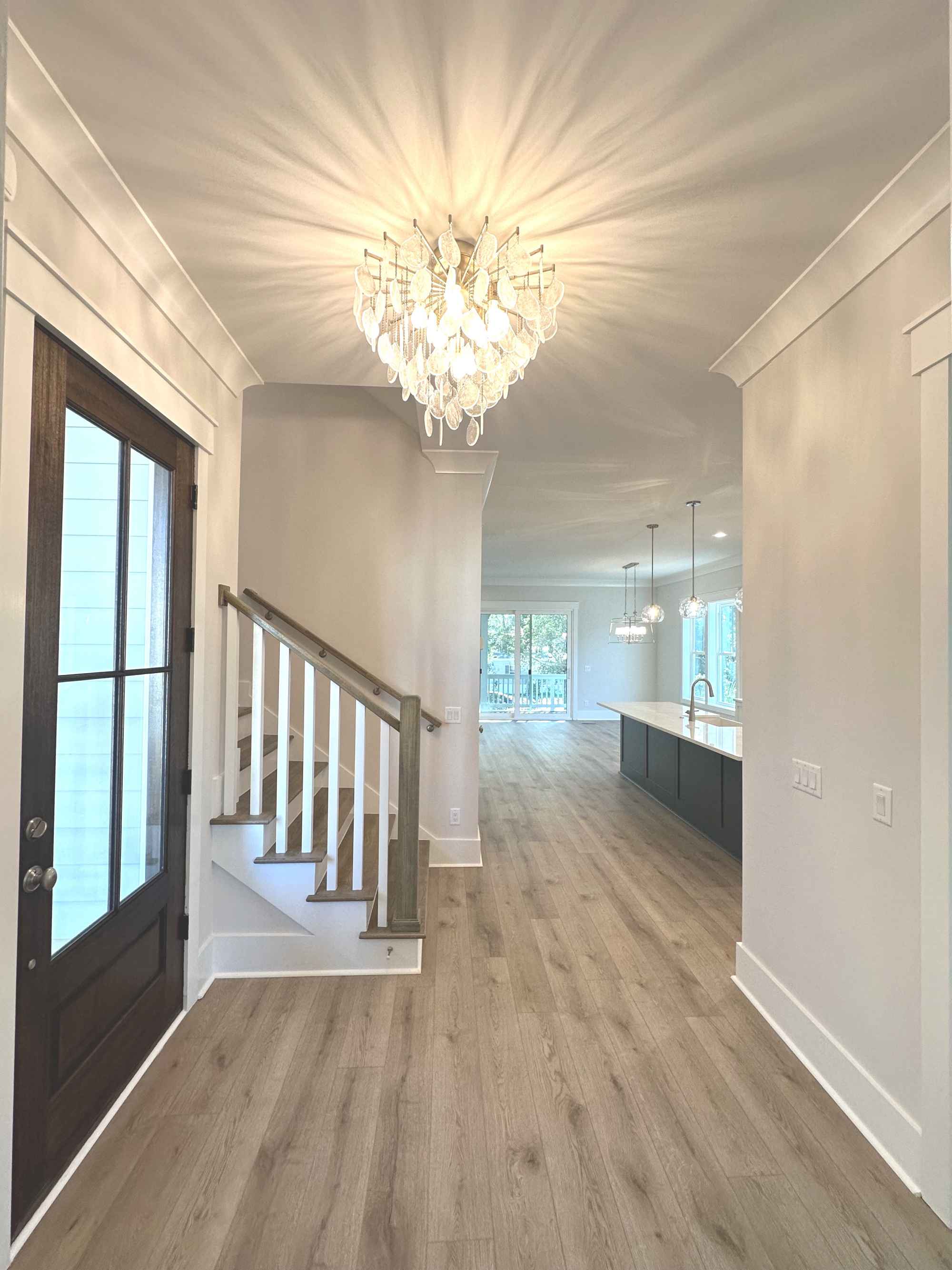 Bright hallway with wooden floors, a modern chandelier, and a staircase on the left. Leads to a spacious kitchen with hanging lights and an airy vibe.