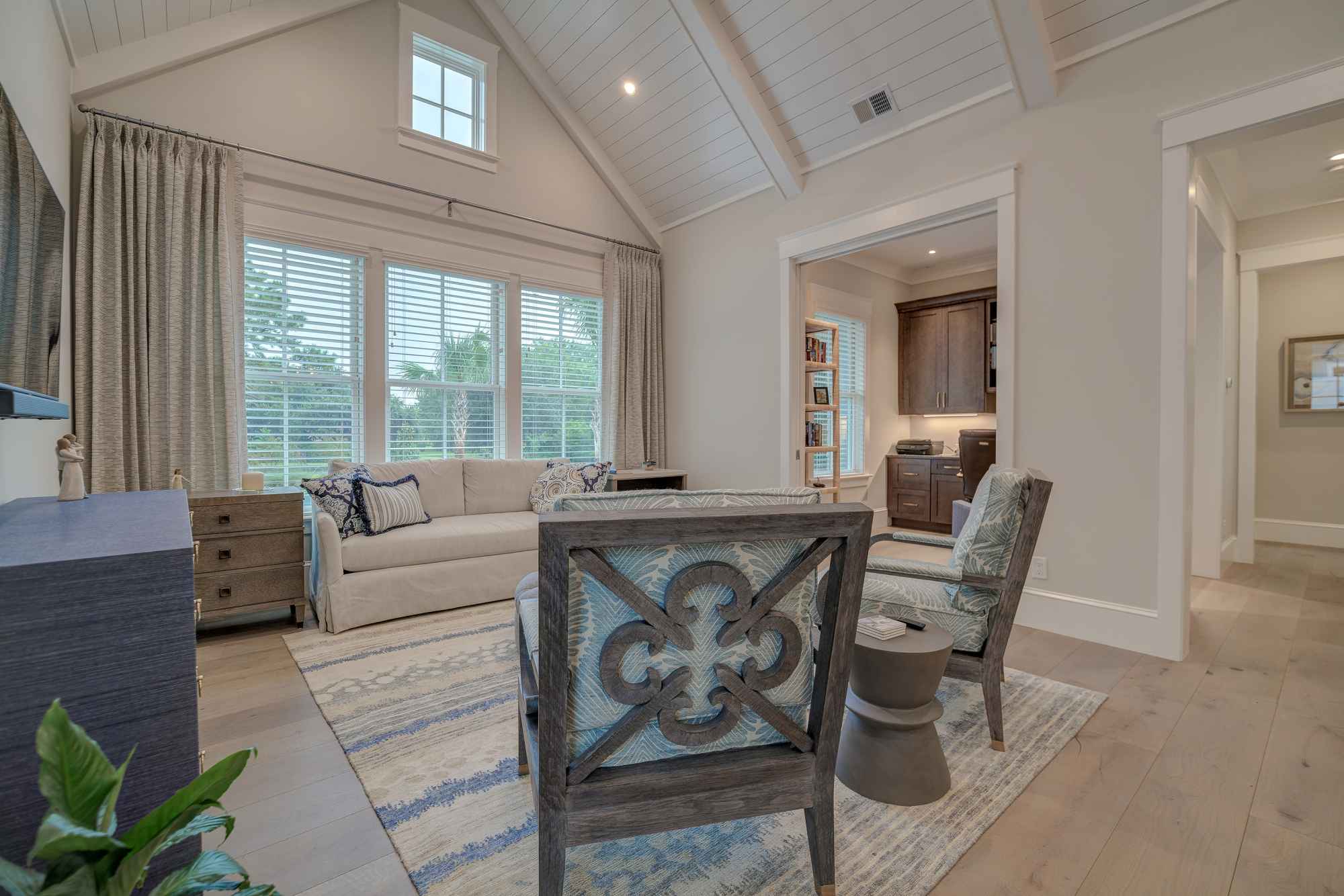 A bright, modern living room with a vaulted ceiling, large windows, comfortable seating, and an adjoining office area.