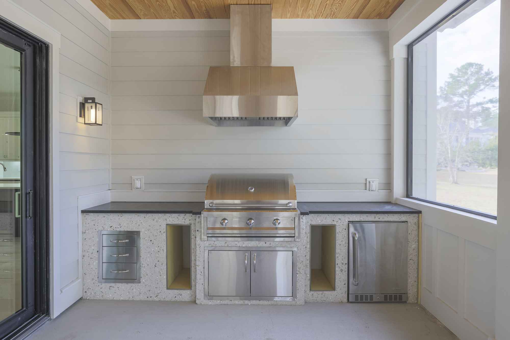 Outdoor kitchen with a stainless steel grill, vent hood, and sleek countertops. Modern design, bright lighting, and a view of the trees outside.