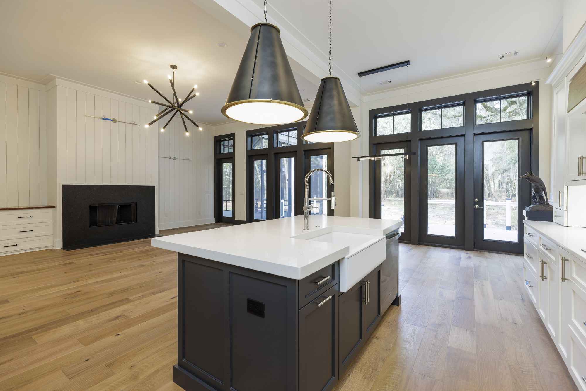 Modern kitchen with hardwood floors, a black island with white countertop, and two large pendant lights. Glass doors offer a view of the outdoors.