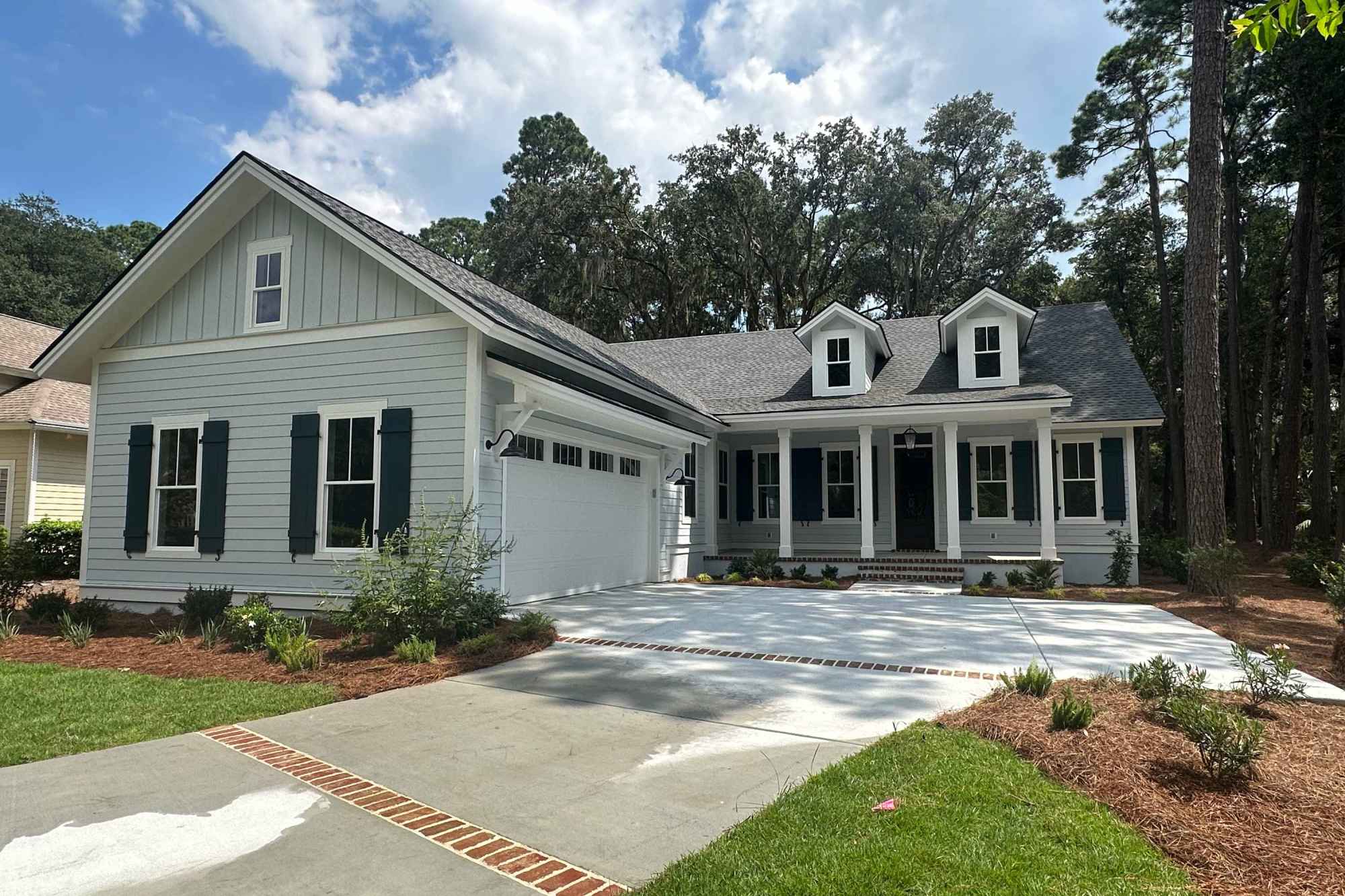 Charming single-story house with light blue siding, dark shutters, and a covered porch. Surrounded by trees and a well-maintained lawn under a blue sky.
