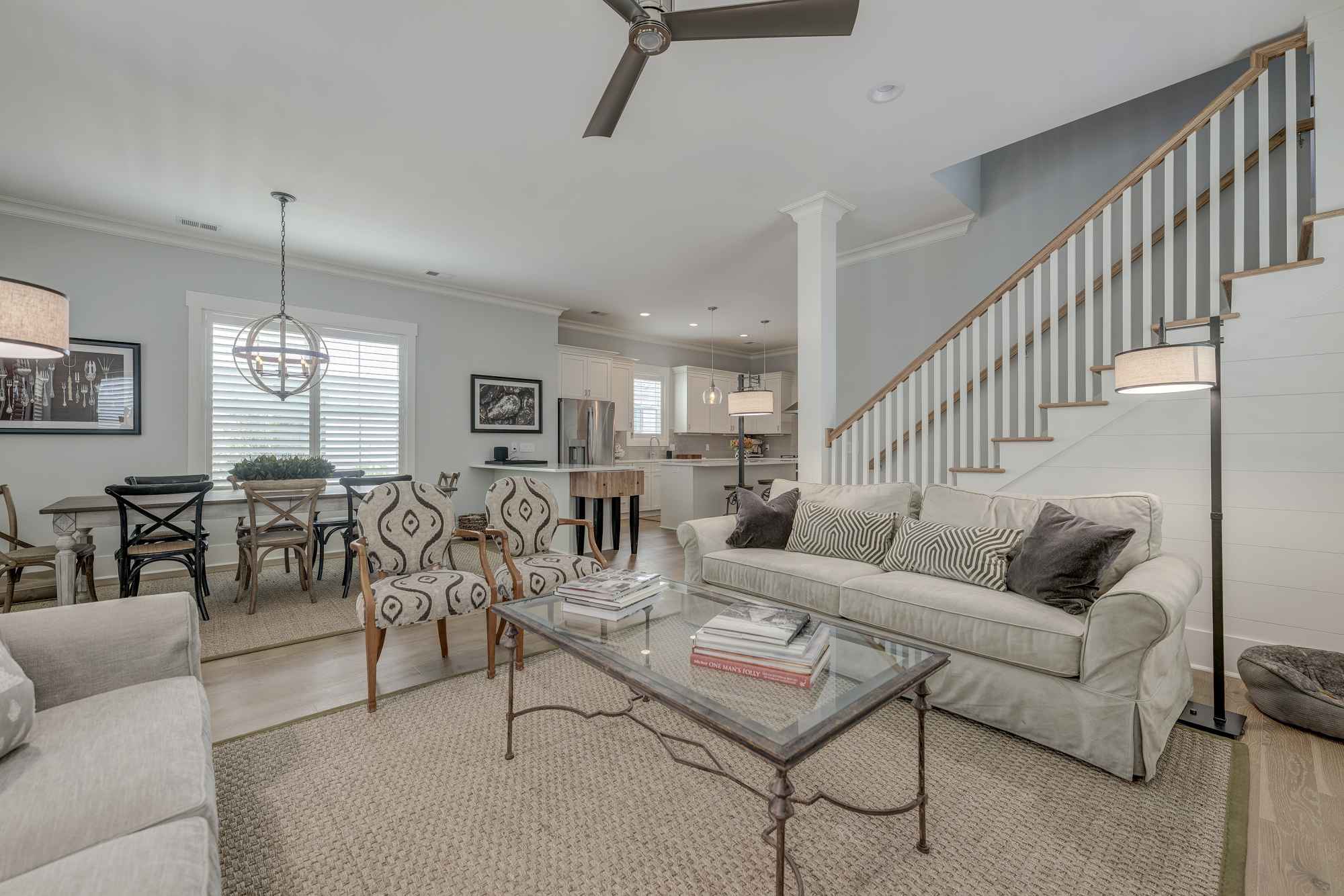 Bright, modern living room with neutral tones featuring two sofas, a glass coffee table, patterned chairs, and a dining area under a stylish pendant light.