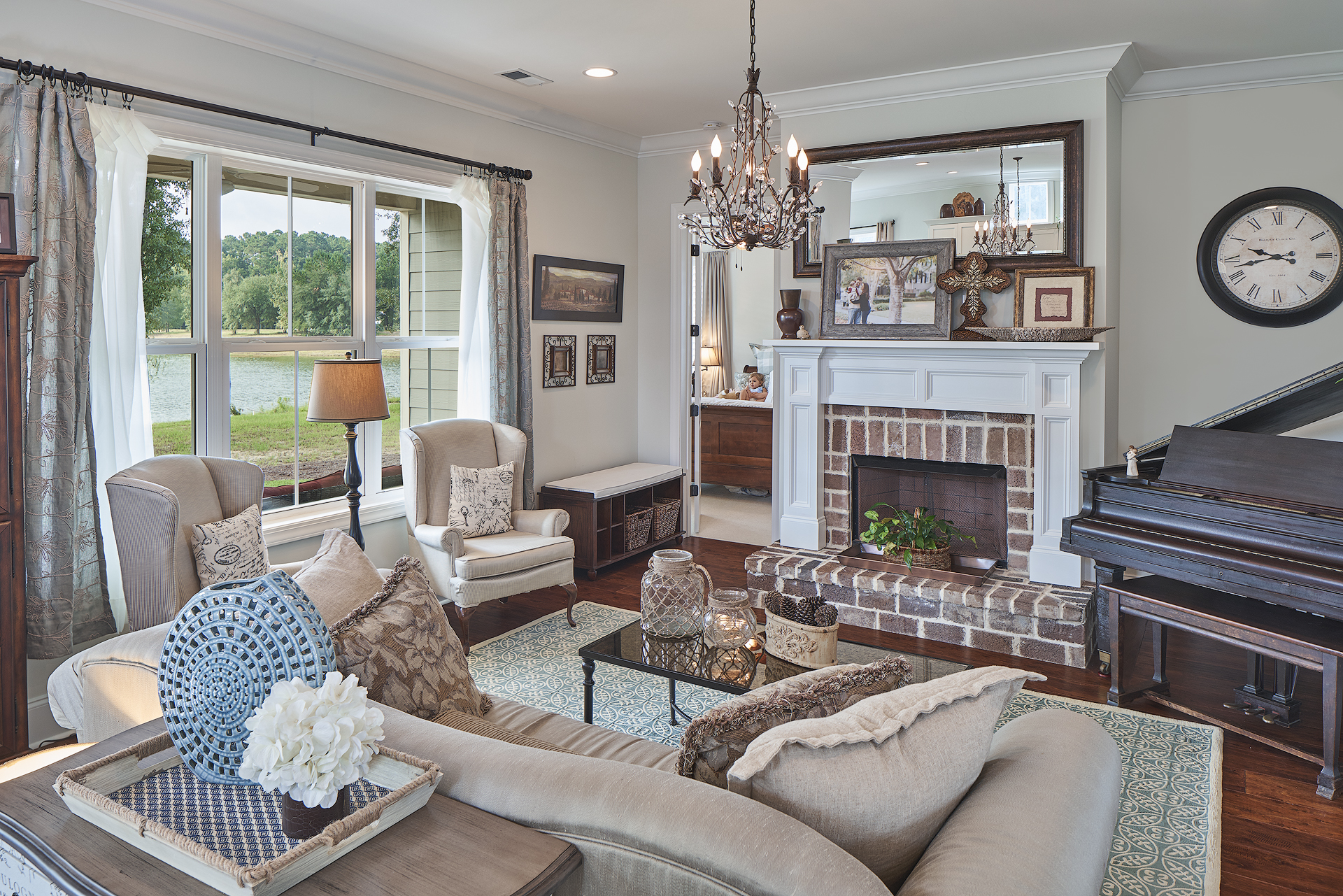 Elegant living room with light furniture, plush cushions, and a chandelier. A grand piano, brick fireplace and large windows with a scenic view add classic charm.