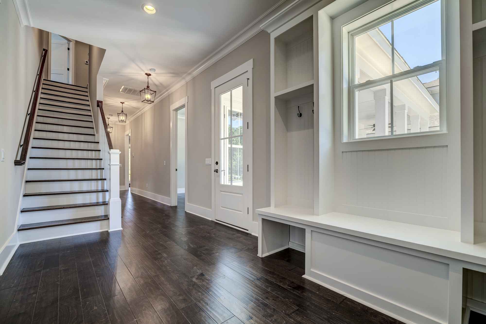 Elegant hallway featuring dark wooden floors, a white staircase with a wooden railing, and bright natural light from large windows.