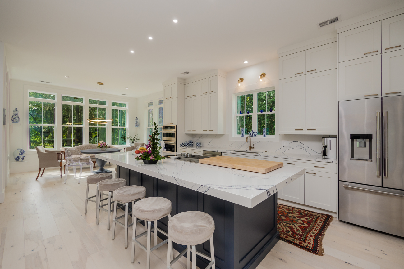 A bright and modern kitchen features a large island with seating, white cabinets and stainless steel appliances. The space opens up to a dining area with large windows overlooking greenery.
