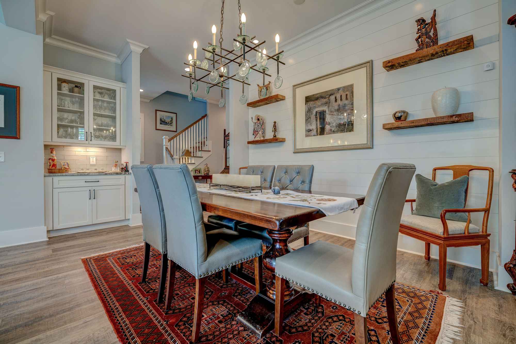 A cozy dining room features a wooden table with gray cushioned chairs on a red patterned rug. A chandelier hangs above, with art and shelves on the walls.