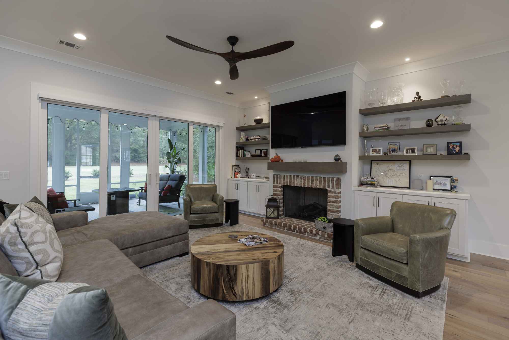 Modern living room with a sectional sofa, two armchairs, and a wooden coffee table. A TV is above a brick fireplace, and large windows offer garden views.