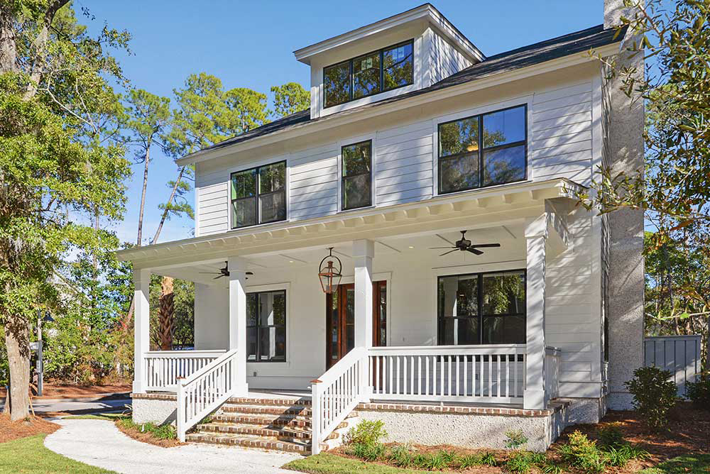 A two-story, white modern farmhouse with large windows, a covered front porch with ceiling fans, surrounded by trees.